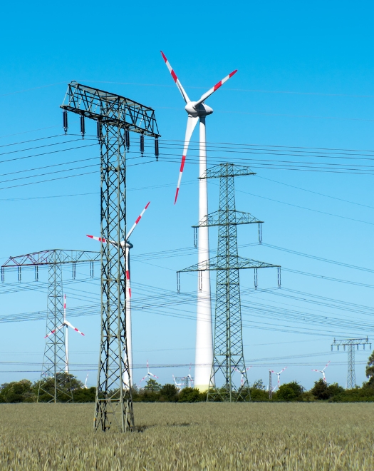 Elektrické vedenie a veterné turbíny na poli pod modrou oblohou – kombinácia tradičnej a obnoviteľnej energie.