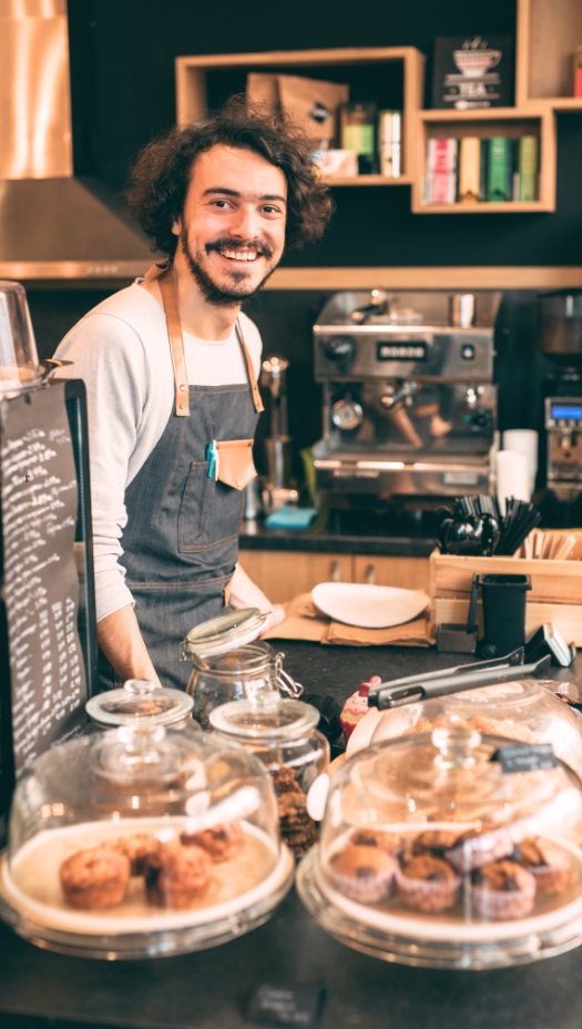 Usmiaty barista v zástere stojí za pultom v kaviarni s vystavenými zákuskami a kávovarom v pozadí. Elgas