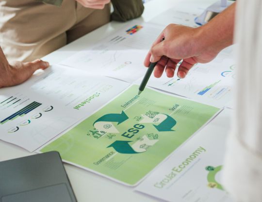 Ruky nad pracovným stolom plným dokumentov s grafmi a zelenou infografikou s nápisom ESG, znázorňujúcou environmentálne, sociálne a riadiace aspekty. Elgas